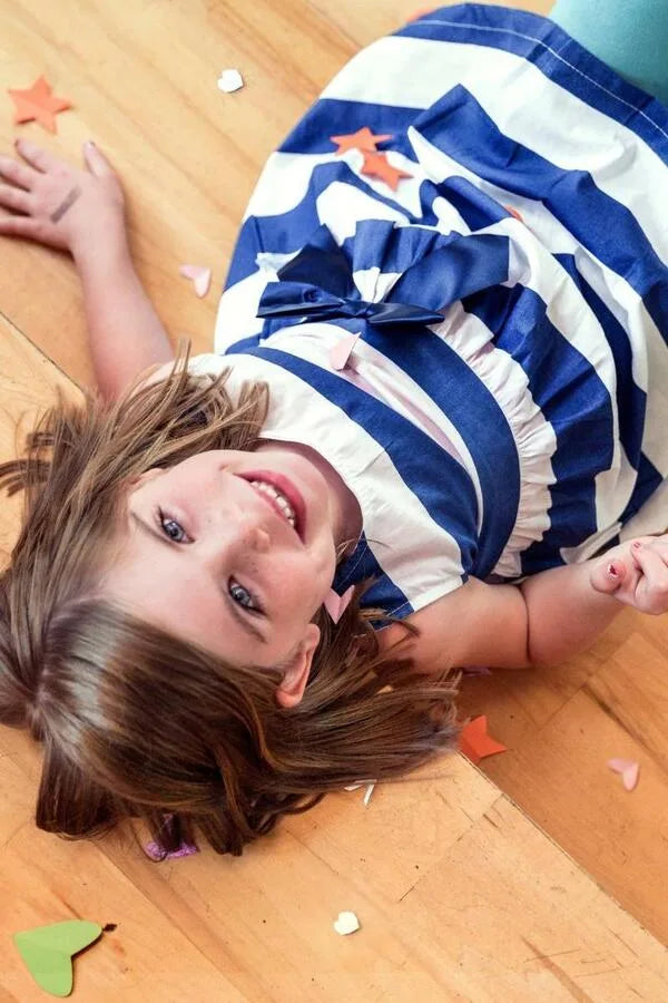 Una ragazzina con un vestito a righe blu e bianche è sdraiata su un pavimento di legno, sorridendo alla macchina fotografica con stelle e cuori di carta sparsi intorno a lei