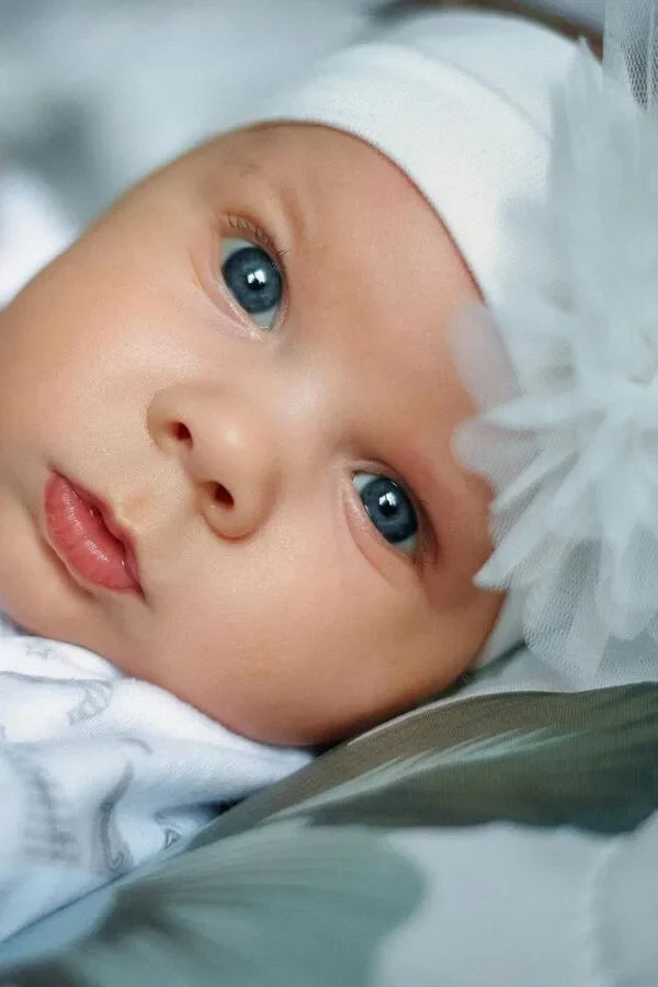 Un primo piano cattura il volto di una neonata con splendidi occhi azzurri. La bambina indossa un cappellino bianco ornato da un delicato fiore bianco.