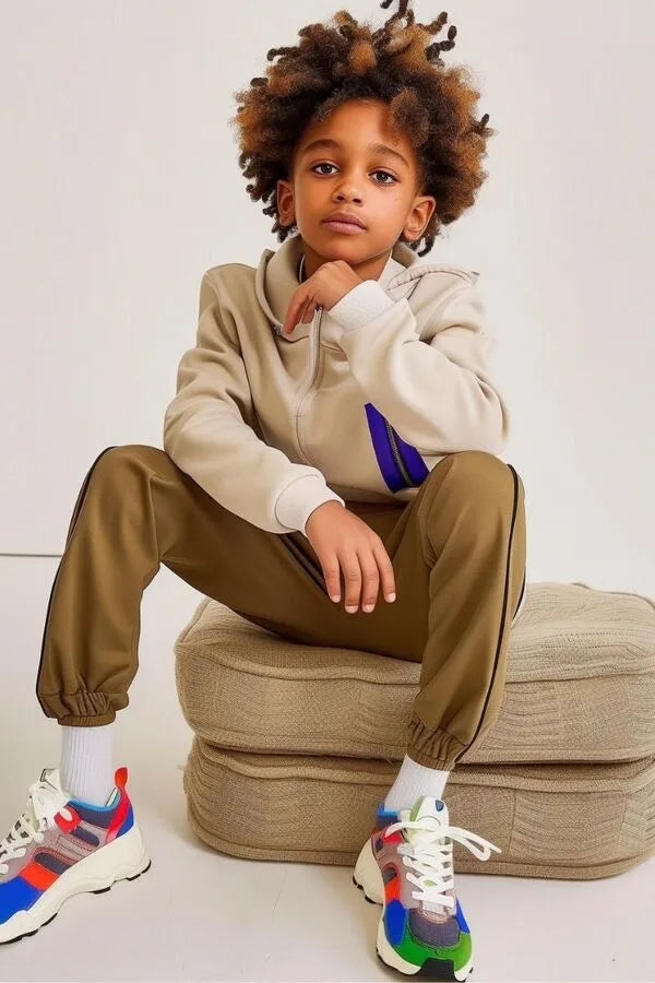 Un giovane ragazzo con capelli ricci e un'espressione pensierosa è ritratto seduto in uno studio con uno sfondo neutro, indossando abiti casual alla moda e scarpe sportive colorate.